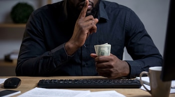 Hombre moreno con barba, vestido con camisa negra, sentado en un escritorio, con un dedo sobre los labios y sujetando un fajo de billetes de cien dólares.