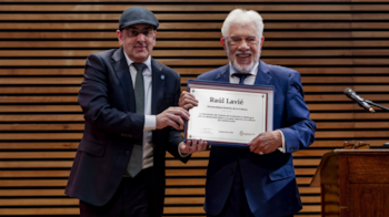 Dos hombres con traje formal sostienen una placa conmemorativa enmarcada ante un panel decorativo de franjas de madera