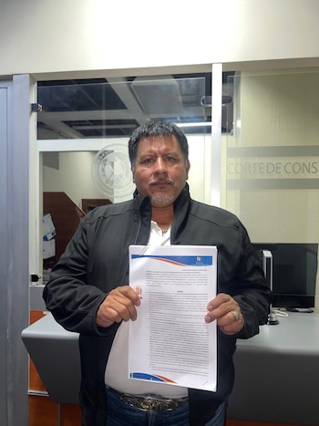 Hombre de cabello oscuro, bigote y barba corta sostiene un documento impreso frente a un panel de vidrio con las palabras "Corte de Constitucionalidad" visibles