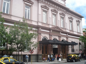 Fachada del Hospital Italiano de Buenos Aires, edificio rosa pálido con detalles marrones, ventanas y marquesina de hierro forjado. Personas caminan y un taxi amarillo