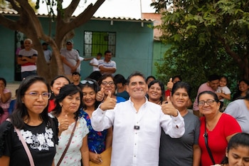 Un hombre sonriente en camisa blanca con dos pulgares arriba está rodeado de una multitud de personas sonrientes, la mayoría mujeres, en un espacio exterior