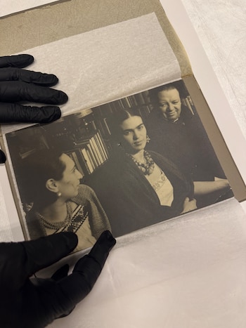 Manos con guantes negros sostienen una fotografía en blanco y negro con tres personas dentro de un libro con papel protector