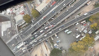 Vista aérea de una autopista de varios carriles con tráfico, un accidente y vehículos de emergencia. Se observan edificios y árboles adyacentes a la vía