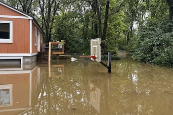 Vivienda de fachada naranja con número 2519, porche y escaleras. Agua turbia cubre el suelo y un aro de baloncesto sumergido. Árboles al fondo