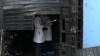 Puerta metálica enrollable semiabierta, dos personas con batas blancas, mascarillas y calzado azul en el interior, reja de metal