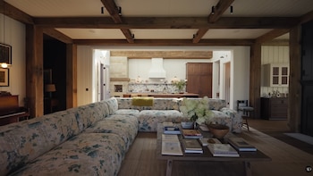 Una sala de estar con sofá floral grande, mesa de centro con libros, vigas de madera expuestas y una cocina moderna visible al fondo