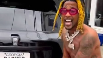 Un hombre sin camisa con rastas amarillas, gafas rojas y collares metálicos posa junto a la parte trasera de un automóvil negro