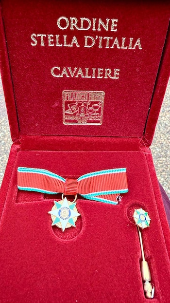 Caja de terciopelo rojo abierta mostrando una medalla de la Orden de la Estrella de Italia con cinta tricolor y una estrella esmaltada, junto a un pin de solapa a juego