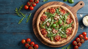 Pizza con masa integral, tomates cherry, rúcula, albahaca y queso rallado sobre tabla de madera. Al lado, tomates frescos y un cuenco con queso en mesa azul.