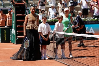 Dos mujeres y dos niños posan sonrientes en una cancha de tenis de arcilla. Las mujeres sostienen raquetas de tenis. La multitud se sienta en las gradas al fondo