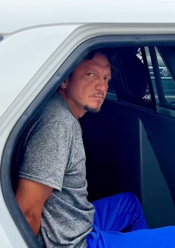Un hombre con camisa gris y pantalón azul está sentado con las manos esposadas en el asiento trasero de un coche blanco con la puerta abierta