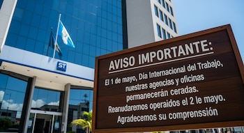 Un edificio moderno de banco con fachada de cristal azul y una bandera de Guatemala, junto a un cartel de madera que anuncia el cierre por el 1 de mayo.
