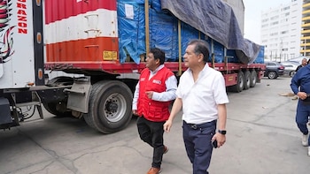 Dos hombres caminan junto a un camión rojo y blanco que transporta carga cubierta con lona azul en un patio exterior, con edificios al fondo