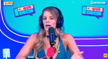 Mujer rubia con auriculares y micrófono en primer plano, fondo azul con letrero "Sin Que Decir" y número telefónico. La mujer usa un top de mezclilla