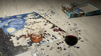 Vista aérea de un rompecabezas incompleto de 'La Noche Estrellada' sobre un piso de madera, con piezas dispersas y dos copas de vino al frente, junto a la caja abierta del puzzle.