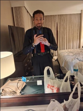 Vince Staples de pie en un espejo, tomándose una selfie con un teléfono. Viste una camisa negra y corbata roja en una habitación de hotel con cortinas y una cama