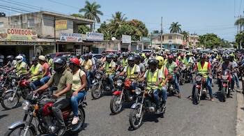 Decenas de personas con cascos y chalecos reflectantes conducen motocicletas por una avenida urbana con edificios, negocios y palmeras bajo el sol.