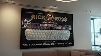 Un gran mural negro en una pared blanca muestra el nombre 'RICK RO$$', álbumes y muchos discos plateados, con '40,500,000 RIAA CERTIFICATIONS' abajo