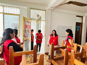 Varias personas, la mayoría con chalecos rojos, interactúan en una sala. Hay sillas de madera apiladas en primer plano, una pizarra blanca al fondo y ventanas