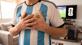Hombre con camiseta de fútbol de Argentina, con el logo AFA y tres estrellas, se toma el pecho. Al fondo, un televisor muestra el logo FIFA World Cup 2026.