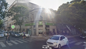 Esquina urbana con una sucursal bancaria de marquesina verde, automóviles en la calle y peatones en el cruce peatonal
