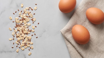 Copos de avena y semillas de lino en el lado izquierdo y tres huevos marrones junto a un paño de lino a la derecha.