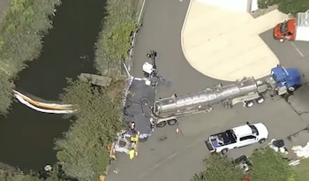 Vista aérea de un camión cisterna plateado y una barrera de contención flotante desplegada en un canal junto a una zona arbolada