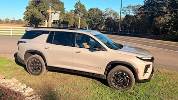 Camioneta Chevrolet Traverse de color beige estacionada sobre el pasto junto a una carretera rural bordeada por árboles y una valla blanca
