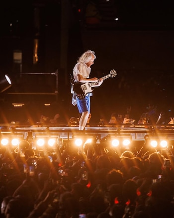 Un guitarrista con cabello largo y camisa blanca toca una guitarra eléctrica oscura en un escenario brillante, con luces doradas y una multitud sosteniendo teléfonos