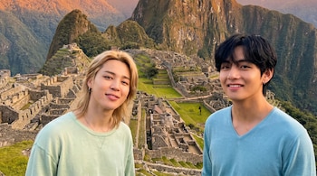 Jimin con cabello rubio largo y V con camiseta azul sonríen frente a la ciudadela de piedra de Machu Picchu y sus montañas.