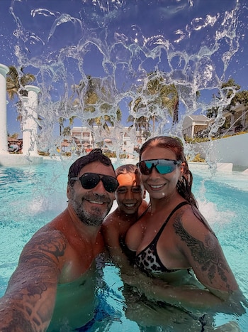 Tres personas con traje de baño y gafas de sol sonríen dentro de una piscina detrás de una cortina de agua que cae