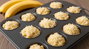 Una bandeja de muffins negra con doce cavidades, cada una llena con masa de color claro cubierta con queso rallado. En el fondo, dos bananas.