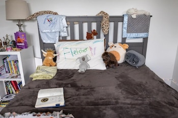 Cama con cubrecama gris, peluches, una almohada escrita con el nombre Fletcher y una estantería blanca al lado con libros y una lámpara