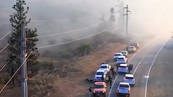 Vista aérea de una carretera rural con fila de vehículos detenidos. Hay personas, árboles, postes de electricidad y cables eléctricos entre el humo
