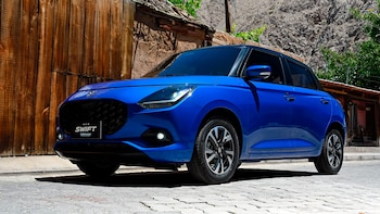 Un automóvil Suzuki Swift Híbrido azul brillante estacionado en una calle empedrada, con una cerca de madera y una pared de piedra al fondo