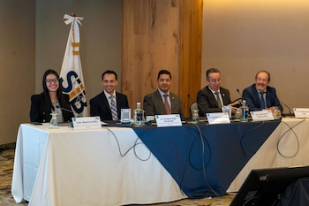 Cinco personas sentadas en una mesa con un mantel blanco y azul, frente a micrófonos y rótulos de nombres, con una bandera blanca con el logo "SBF" de fondo