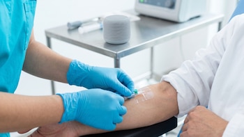 Manos con guantes azules fijan una vía en el brazo de un paciente cerca de una mesa metálica con un altavoz inteligente