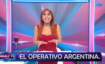 Magaly Medina, presentadora de televisión, con cabello rojizo y vestida de rojo, gesticulando en un estudio de televisión con fondo abstracto azul y rosa