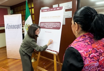 Una mujer de cabello oscuro, vestida de verde, firma un gran póster blanco en un atril. Detrás, una bandera mexicana y un cartel de FONACOT