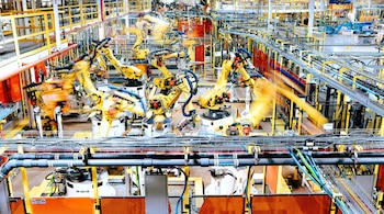 Múltiples robots industriales amarillos con brazos articulados y en movimiento operan en una línea de producción dentro de una fábrica con cintas transportadoras, tuberías y estructuras metálicas