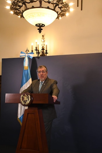 Un hombre con traje y corbata habla en un podio de madera. Detrás, una bandera guatemalteca y un fondo azul oscuro. Arriba, una lámpara de techo y un candelabro
