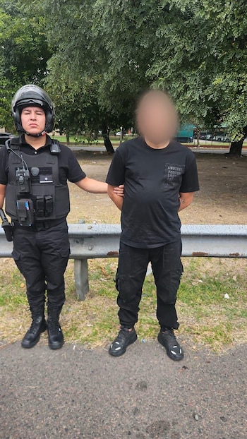 Oficial de policía con uniforme negro y casco, detiene a un hombre vestido de negro con las manos atrás, junto a una barandilla. Hay árboles y una calle detrás