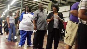 Varias personas de pie en una fila dentro de un edificio. Una mujer con chaqueta negra y blusa de rayas habla y gesticula. Un hombre con camisa gris la observa. Otros individuos están cerca