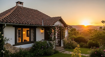 Vista frontal de una casa blanca con techo de tejas rojas y un jardín con rosas rosadas, iluminada por el atardecer dorado en el fondo montañoso.