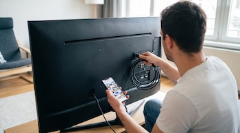 Un hombre conecta un cable HDMI negro a la parte trasera de un televisor. En la otra mano sostiene un teléfono móvil con la pantalla encendida.