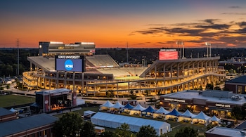 Vista panorámica al atardecer de un estadio iluminado con el logo de la NCAA, rodeado de carpas de eventos, un escenario y comercios.
