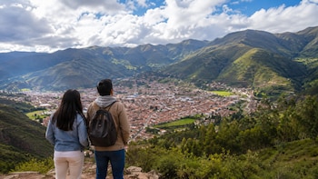 Dos personas de espaldas observan una ciudad con edificaciones de techos rojos en un valle montañoso con vegetación y cielo nublado