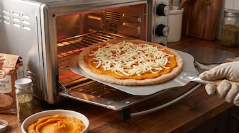 Mano enguantada introduce pizza de calabaza y muzzarella con base integral en horno tostador; hay harina, orégano y puré de calabaza en la mesada.