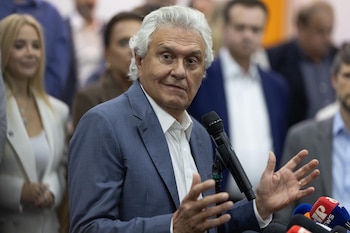 Ronaldo Caiado, un hombre de cabello blanco y traje azul, gesticula con las manos mientras habla en un podio con micrófonos de prensa frente a una audiencia