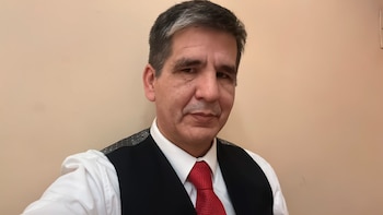 Hombre de mediana edad con cabello oscuro y canoso, viste camisa blanca, chaleco oscuro con puntos claros y corbata roja, sobre un fondo beige liso